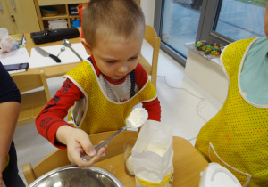 Skupiony Mariuszek przesypuje łyżką mąkę do szklanki.